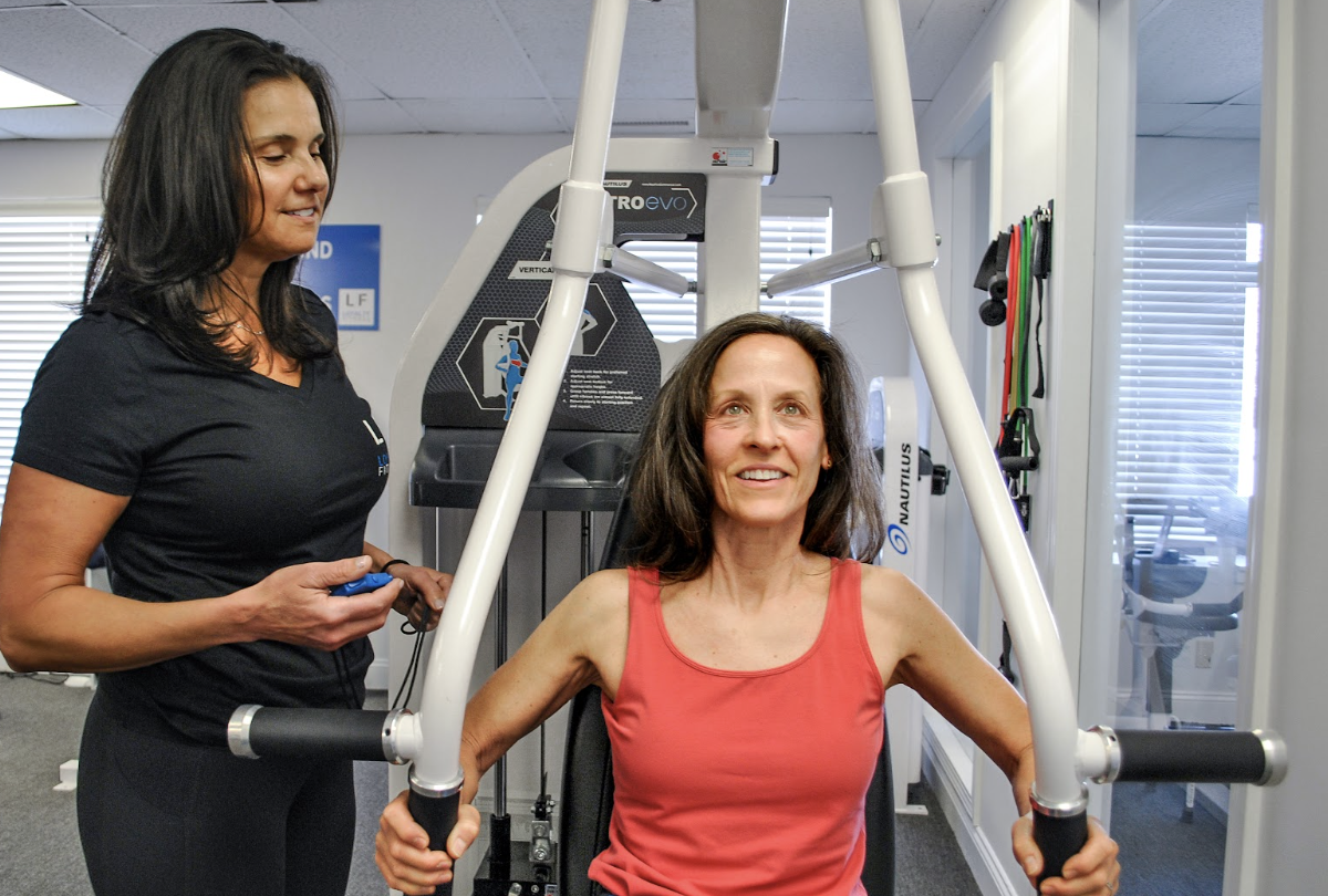 Woman using a workout machine with a trainer beside her.