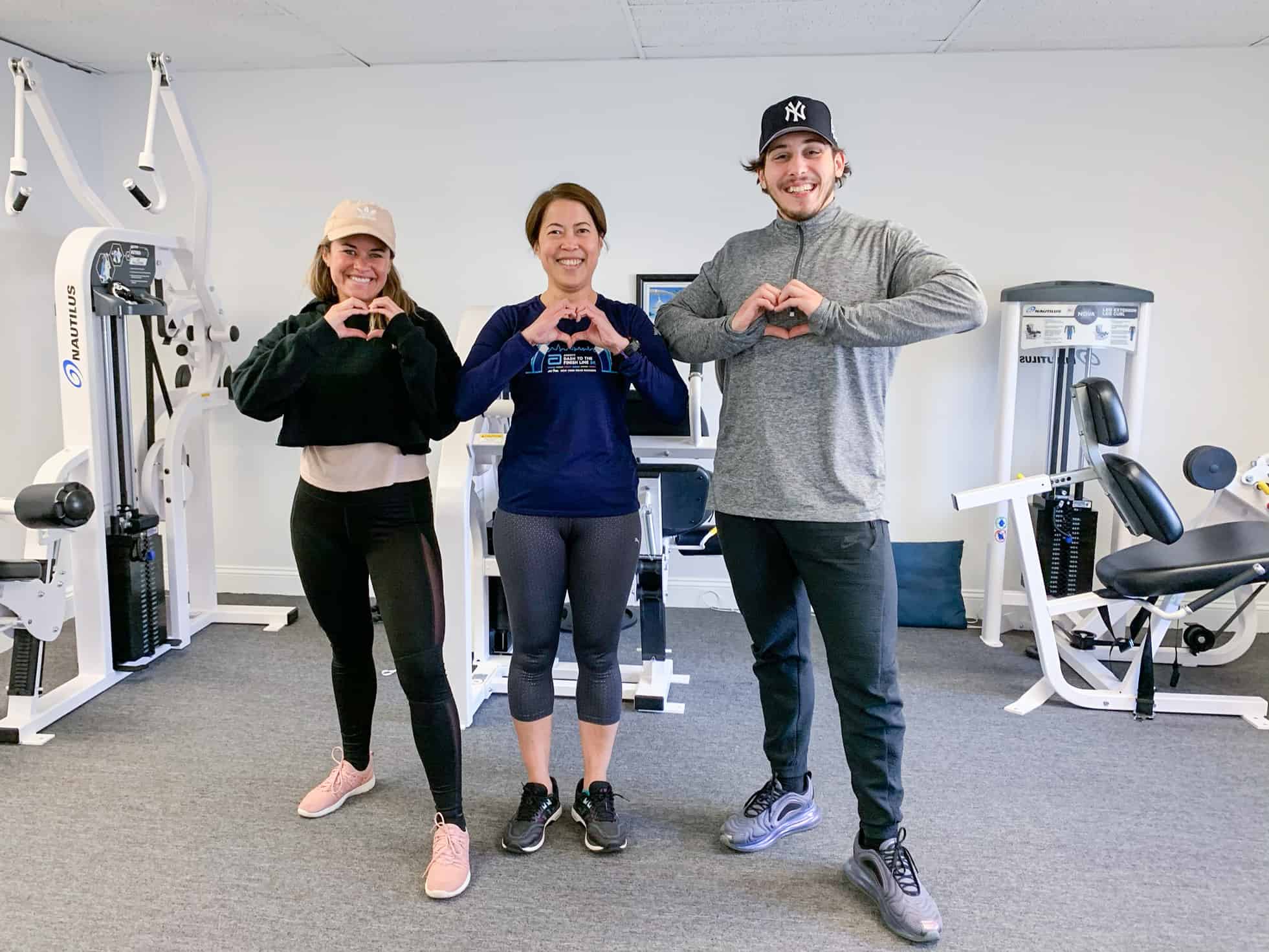 3 Loyalty Fitness trainers in front of equipment with hands in shape of heart.