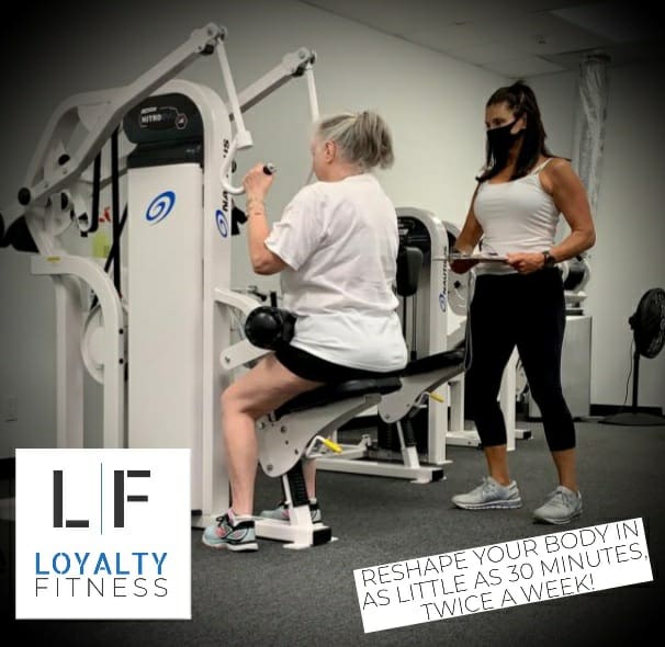 woman using weight machine while working with personal trainer wearing a mask