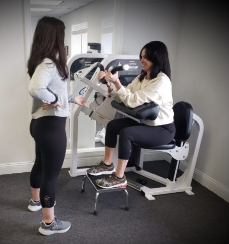 A woman using an arm curl machine while her trainer advises her.