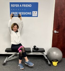 Laura L. holding weight above her head sitting down on bench