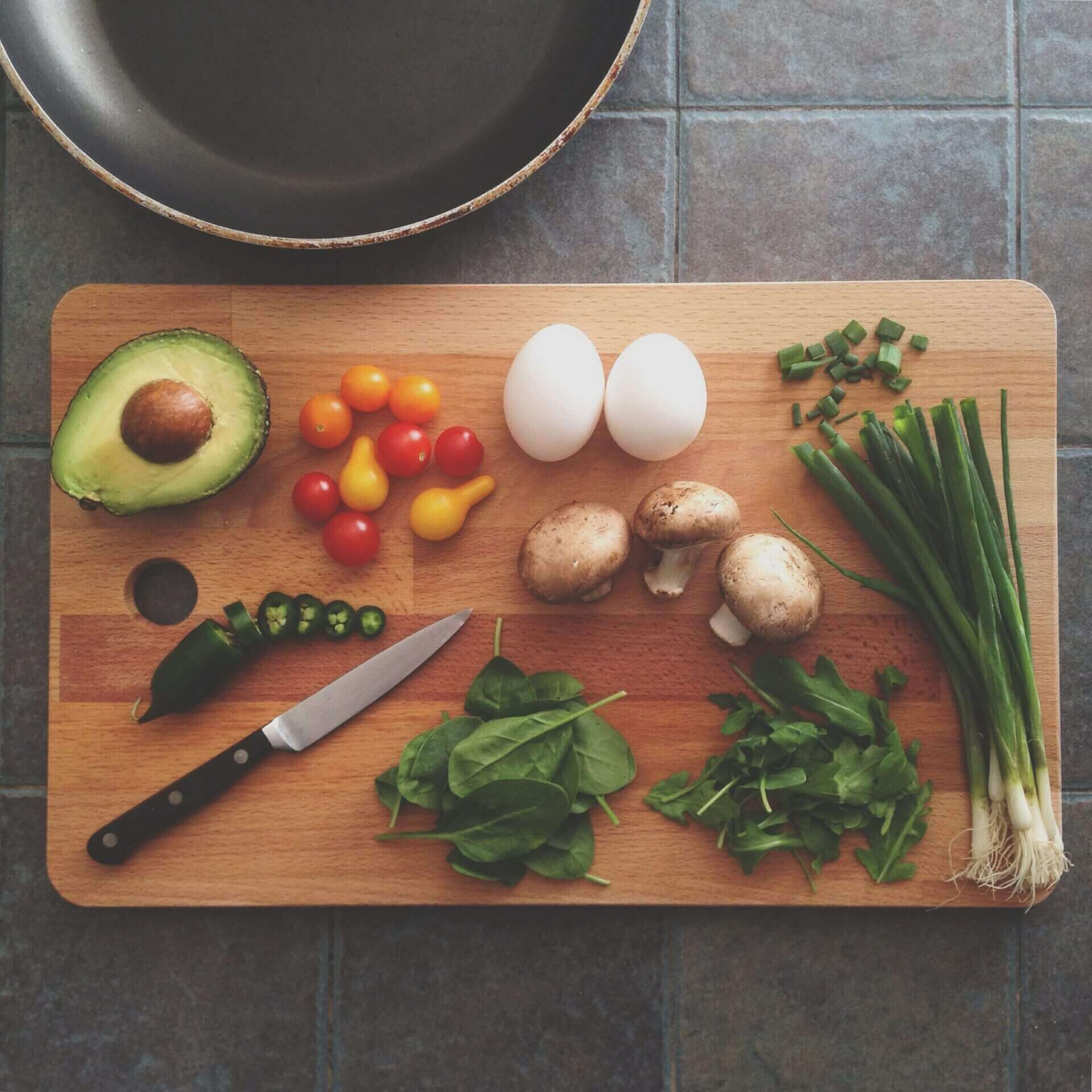Wooden cutting board with eggs, avocado, spinach, mushroom, tomatoes, green onions, and peppers with a knife.