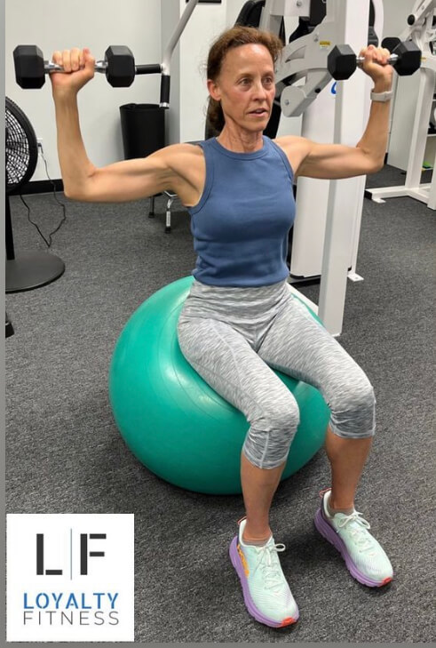 Woman sitting on a aqua yoga bar while lifting weights at an indoor gym.