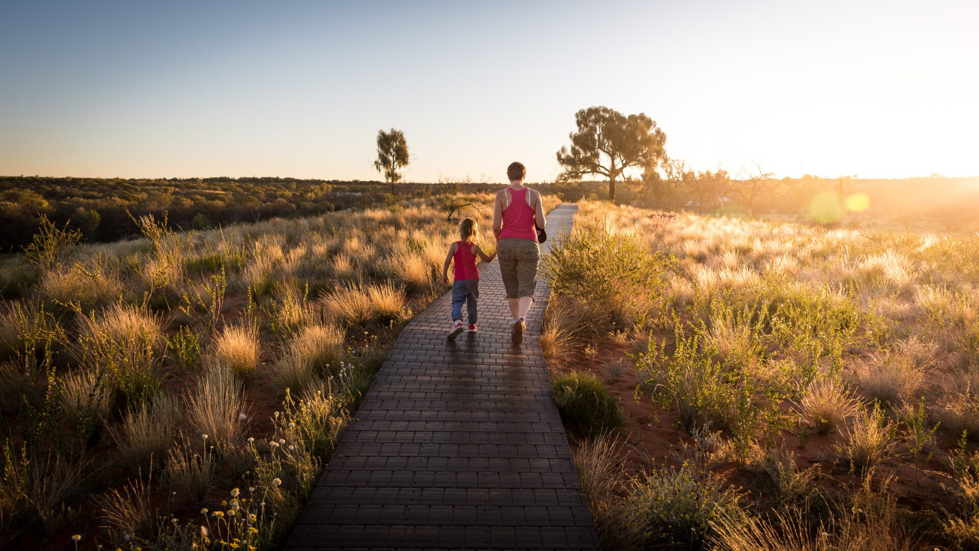 Mom walking with her daughter on a trail with grass on both sides during a sunset.