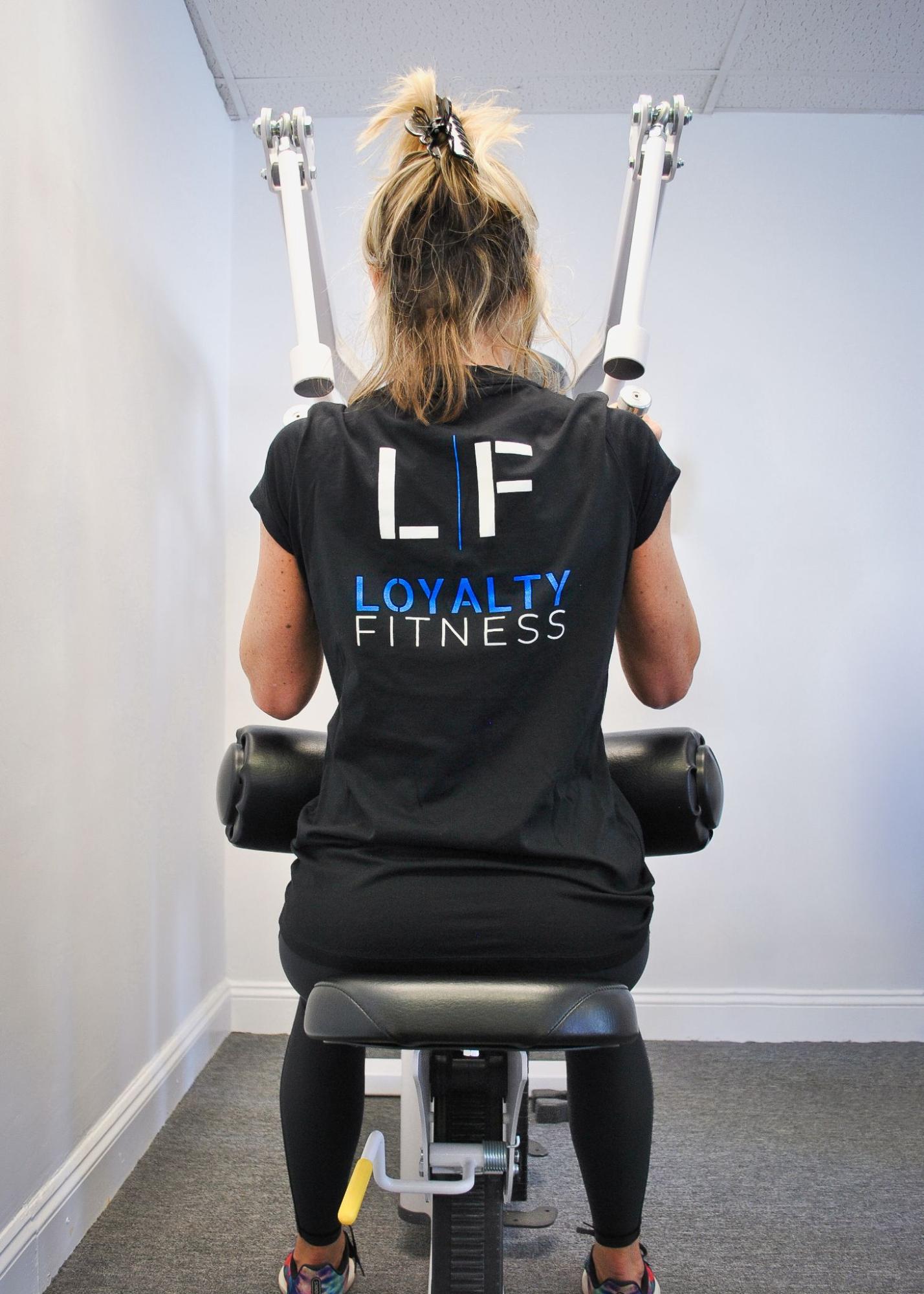 Woman wearing a shirt that says “Loyalty Fitness” with the Loyalty Fitness logo while working out on an exercise machine.